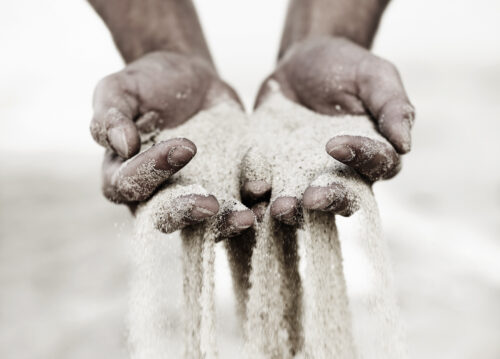 In a sepia-and-white color palette, two hands cup a pile of sand, much of which is falling between the fingers.