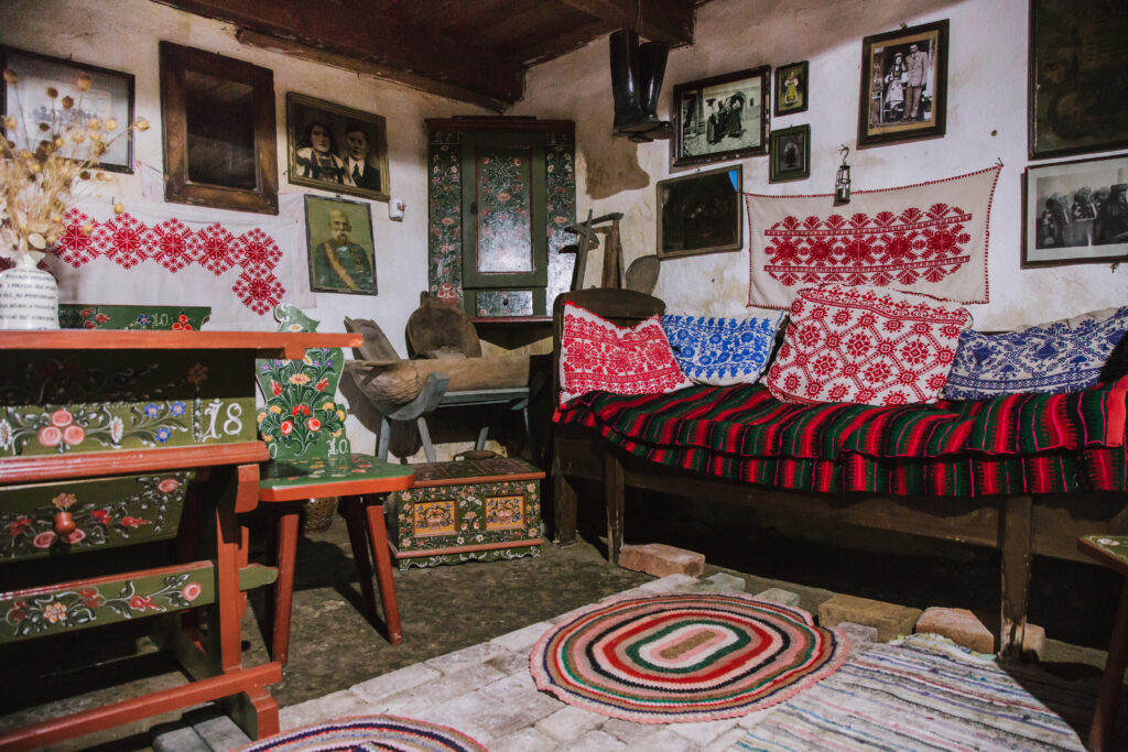 A small room in a house has a wooden cabinet and chair painted green with flowers on one wall, and thin banquette with a colorful blanket and pillows on it on the adjacent well. Pictures, a vase, and hung embroidered cloths cover the walls above.