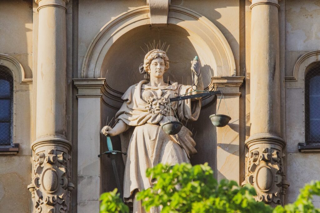 A beige stone statue of a woman wearing a thick robe and holding a gray balancing scale stands under an arch and between two ornately carved pillars.