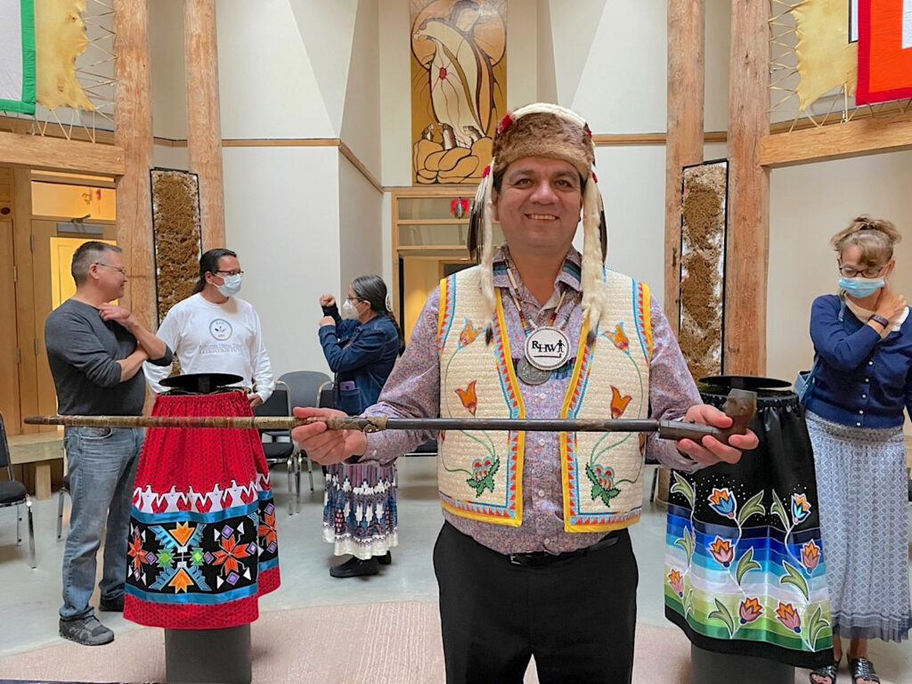 In the image foreground, a person wearing a fur hat with feathers, a medallion and beaded necklace, and an embroidered vest stands in a museum atrium where people chat with one another. The featured person smiles and holds a long pipe horizontally.