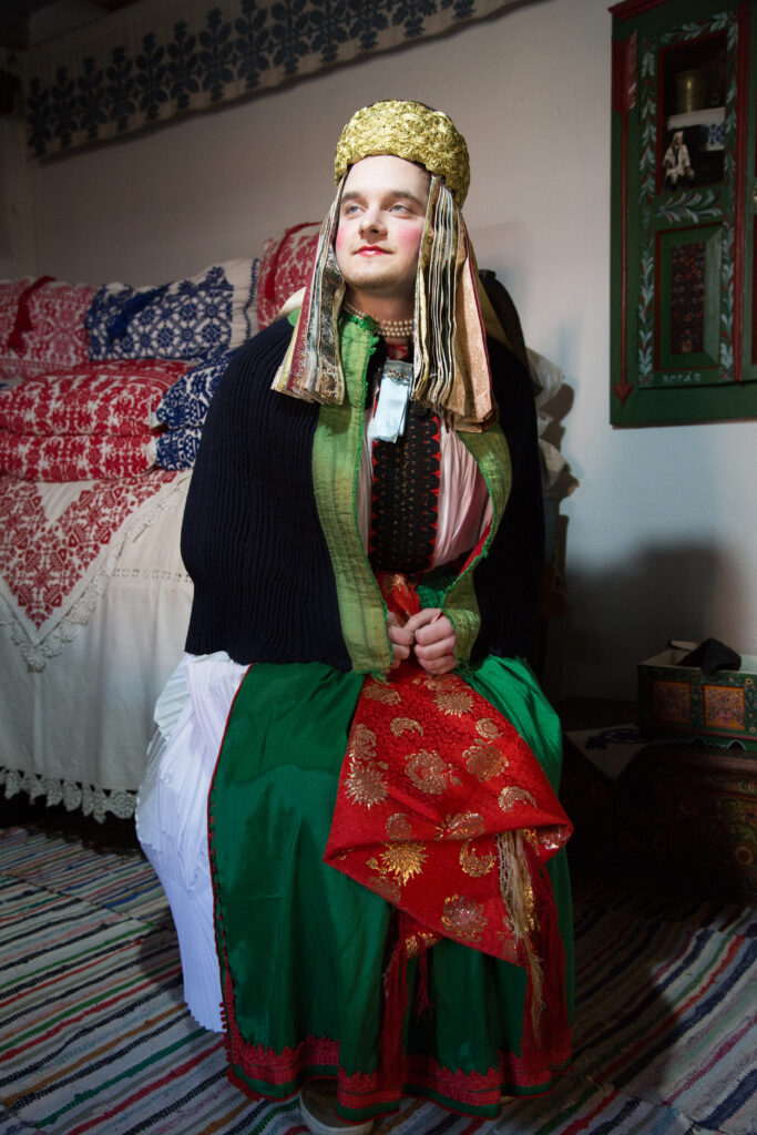 A man in colorful garments with a face made pale and cheeks blushed by makeup sits in a room, posed with his look angled to his right and his body, left. Embroidered pillows and a striped carpet decorate the room.