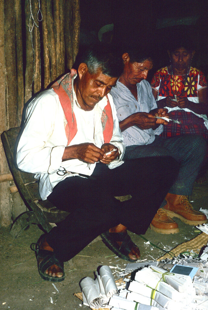 Seated in a row against a wood-slatted wall, each of three people hold and fold paper in front of a large pile of other strips of paper.