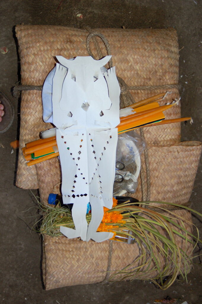 A figure cut into a piece of paper sits on top of several thin wooden instruments and candles, all atop two small bundles woven from palm leaves.