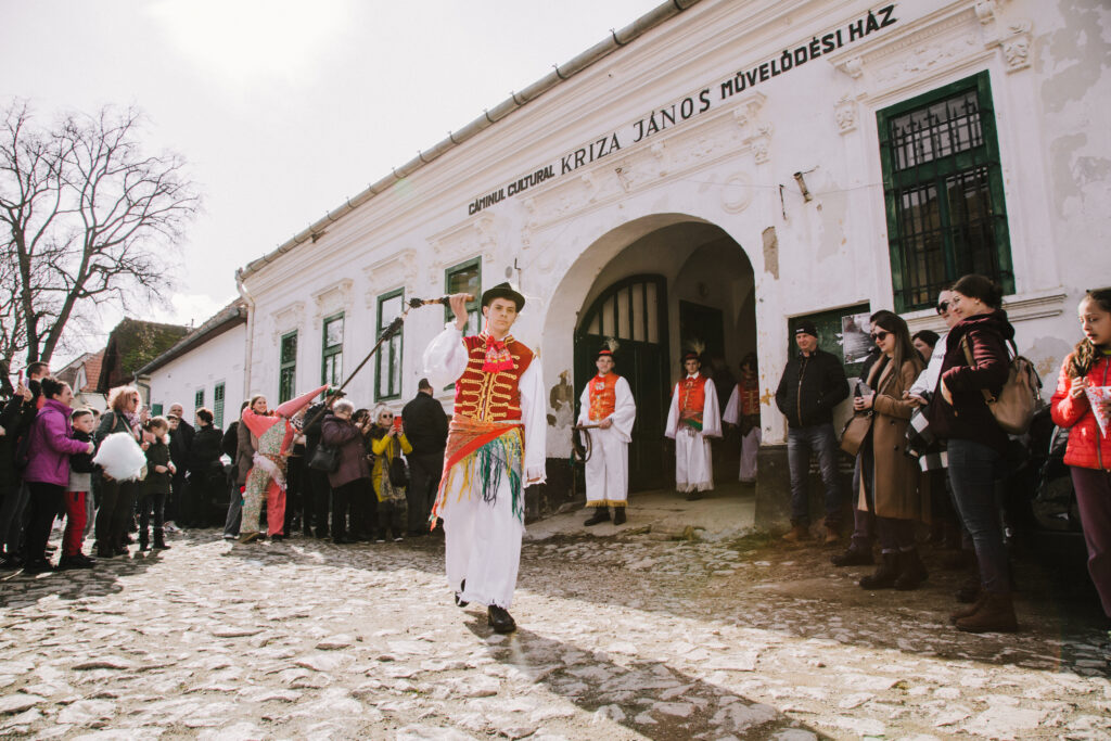 A crowd of people gather in front of a white building as a line of people in white shirts and pants and red vests with gold accents walk through with long items in hand.