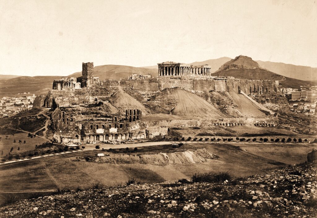 In sepia tones, a landscape has a rocky foreground and tall hills lined with buildings on the horizon. A large, pillared building sits on top of a flat hill, which is lined with a brick wall.