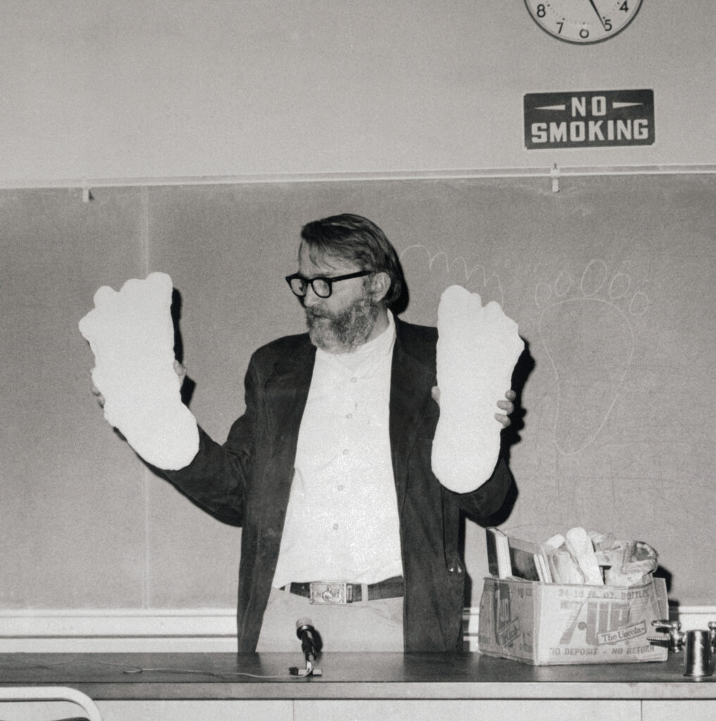 In a black-and-white photograph, a person with a large beard wearing a black suit jacket, white button-down shirt, and khakis stands in front of a chalkboard and holds up two large white objects shaped like footprints.