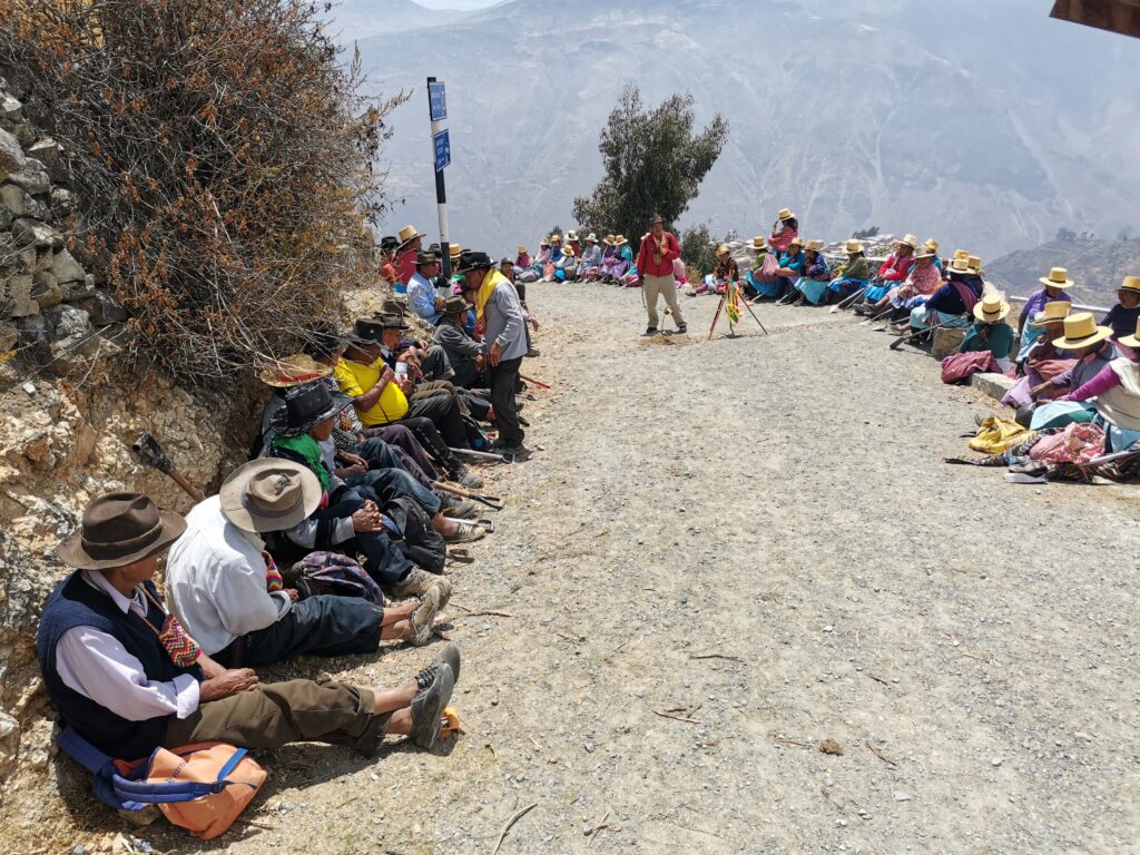 Several people sit along both sides of a gray gravel road with a few people setting up a temporary shrine a display in the road’s center. The road curves in the back of the image and mountains are visible in the distance.