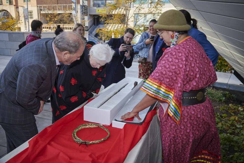 A person wearing colorful clothing and beaded earrings stands on one side of a long table covered with a red table runner. A white box with a long thin object in it sits on top. Two people stand on the other side of the table looking at the object.