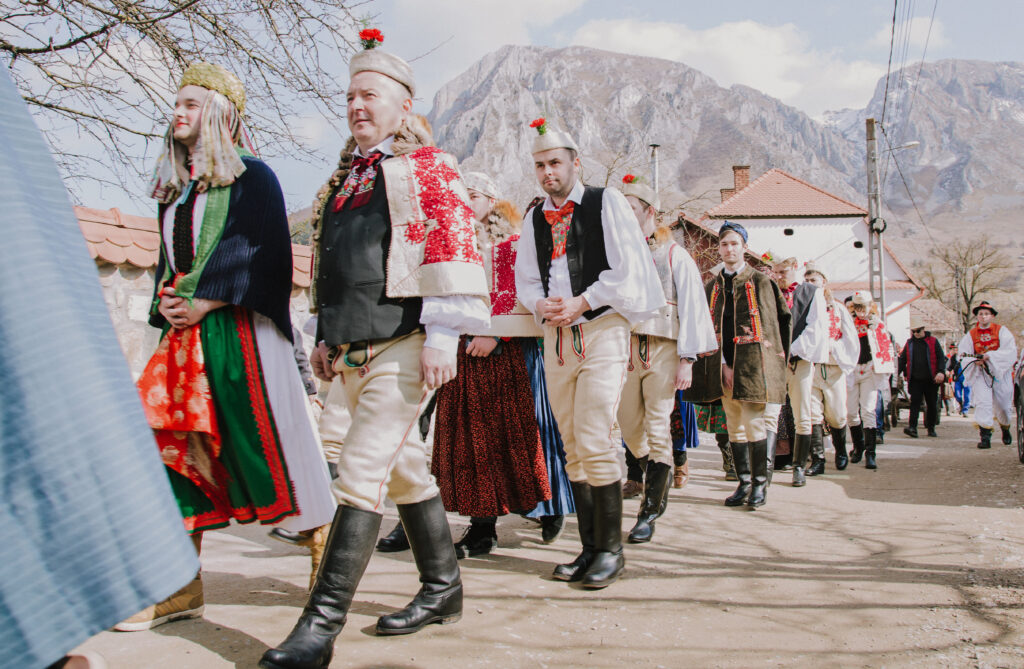 In parallel lines, many people walk in pairs. People wear makeup, dresses, long white curled wigs in one line; and decorated shoulder shawls, beige pants, and hats – each topped with a red flower – in the other line.