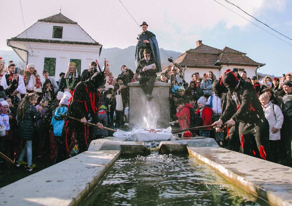 A large crowd of adults and children gather in a public square around a large rectangular stone basin full of water. In the center, masked people dressed in black outfits with red accents use long items to strike a white object laid on the basin’s front edge.