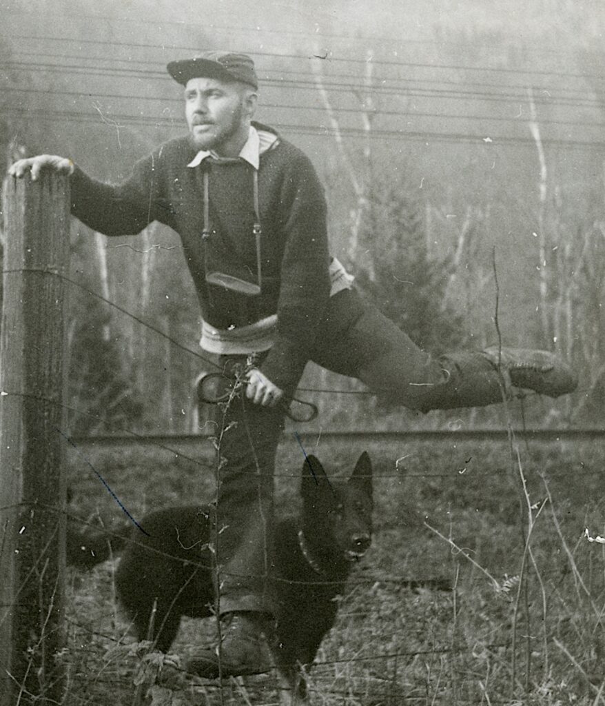 In a black-and-white photograph, a person wearing a cap has a camera hanging around their neck and perches on a wire fence on their right leg. A black dog is on the ground under them.