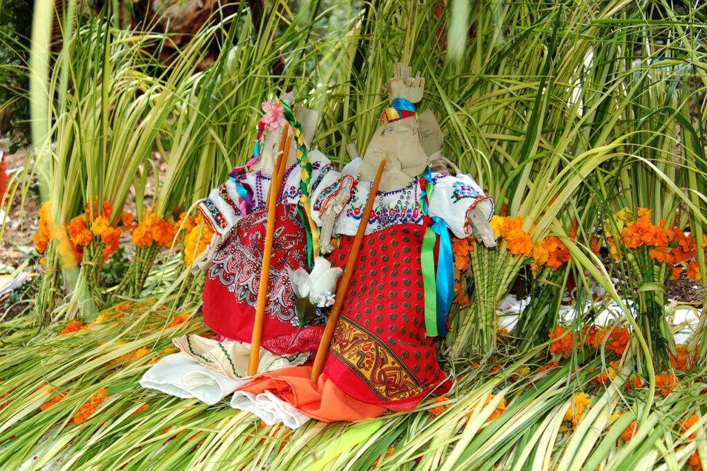 Against and on top of arranged palm leaves and orange marigold flowers sit two paper figure cutouts dressed in colorful ribbons and patterned cloth.