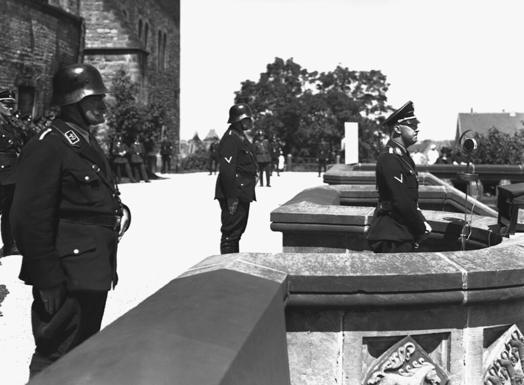 In black and white, people in uniforms and helmets stand behind a single person, also in uniform, who stands on a curved balcony in front of a standing microphone.