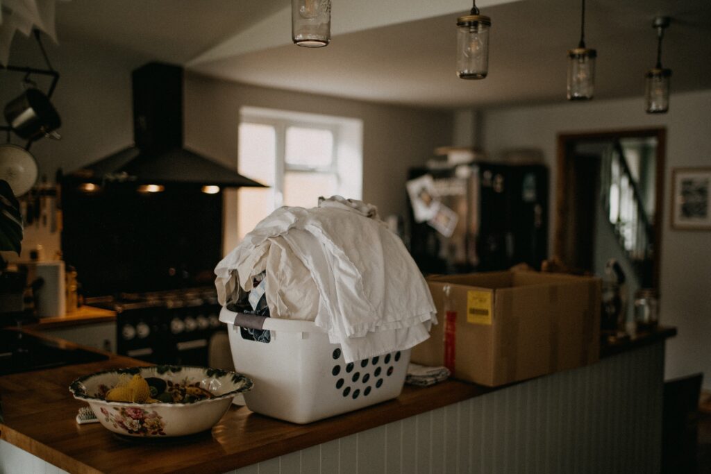 A white laundry basket overflowing with clothes sits on a kitchen counter between a cardboard box and a decorative bowl.