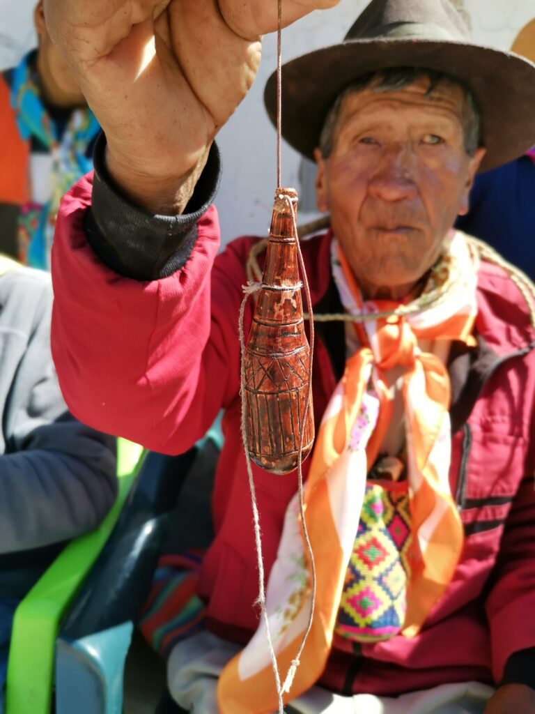 A close-up image centers an oblong wooden object etched with a design. The object has a string tied to it, which is held by an older person with facial wrinkles wearing a brown hat and neck scarf.