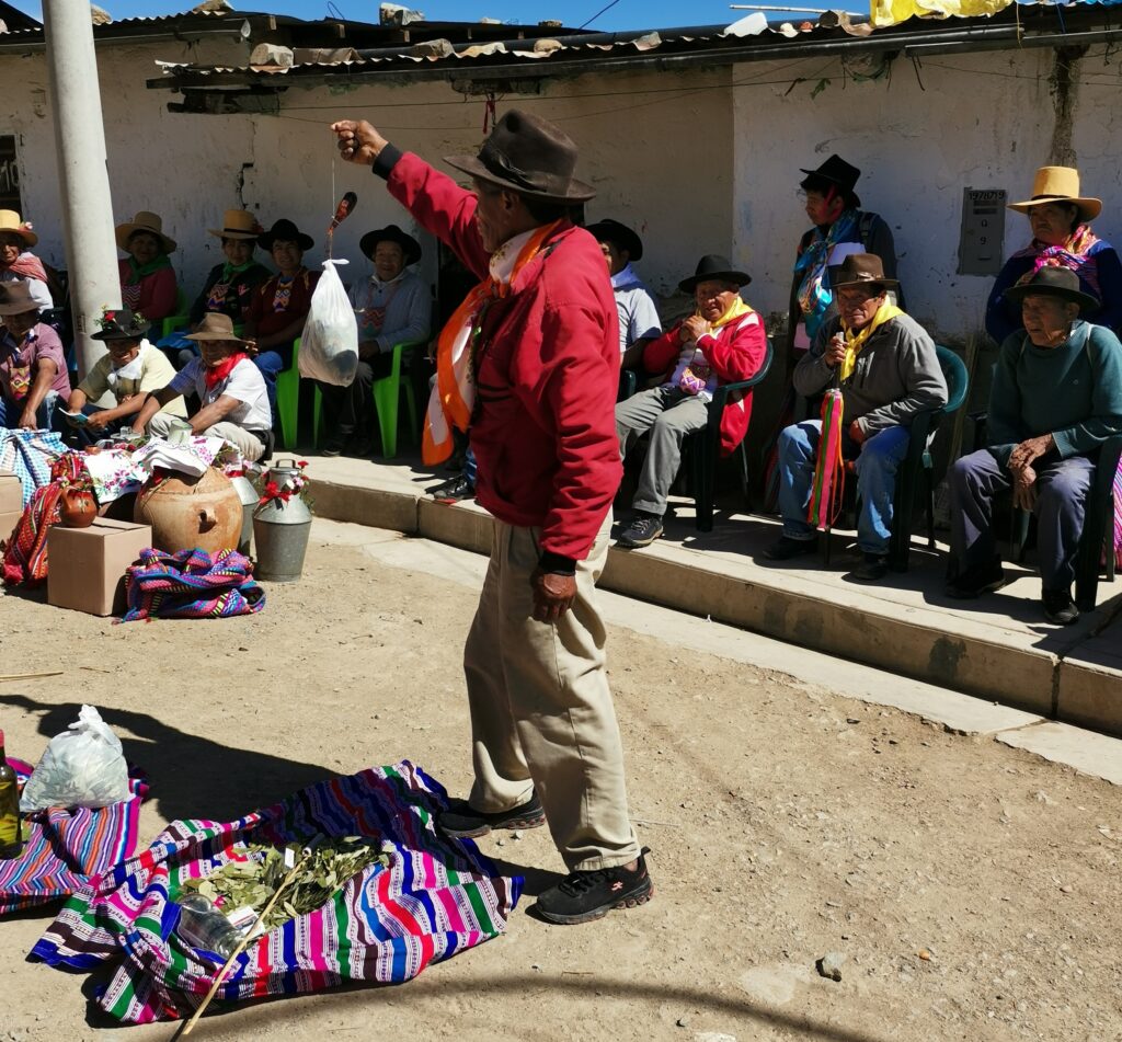 In front of many people seated around the perimeter of a town square, a person in a brown hat stands in its center over a pile of green leaves on a colorful patterned cloth. They are holding up a bag by a string.