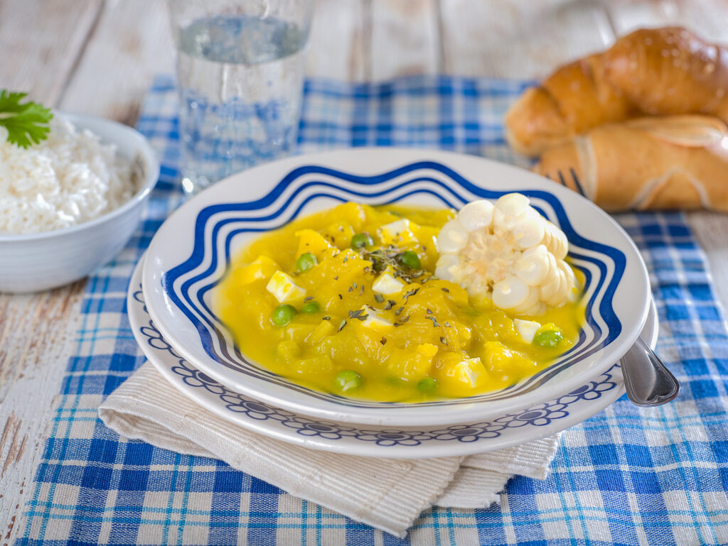 A yellow stew with green peas and a small piece of white corn lies on a white and blue bowl on a white and blue checkered tablecloth with a bowl of rice, two bread rolls, and a glass of water in the background.