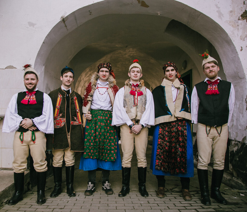 Six people stand in line in front of a stone archway. Three wear beige pants and hats (the latter with a red flower) and tall black boots. The other two wear blue skirts, cloaks, flower-print waist aprons and headwraps, and red lipstick.