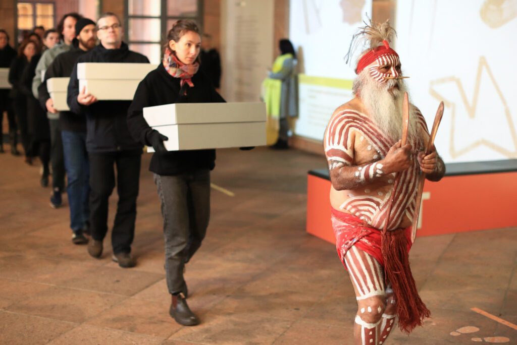 A person with a long gray beard and red and white designs painted on their face, chest, and legs wears a feathered headdress and holds two wooden sticks. They lead a procession of several people in black clothes carrying white cardboard boxes.