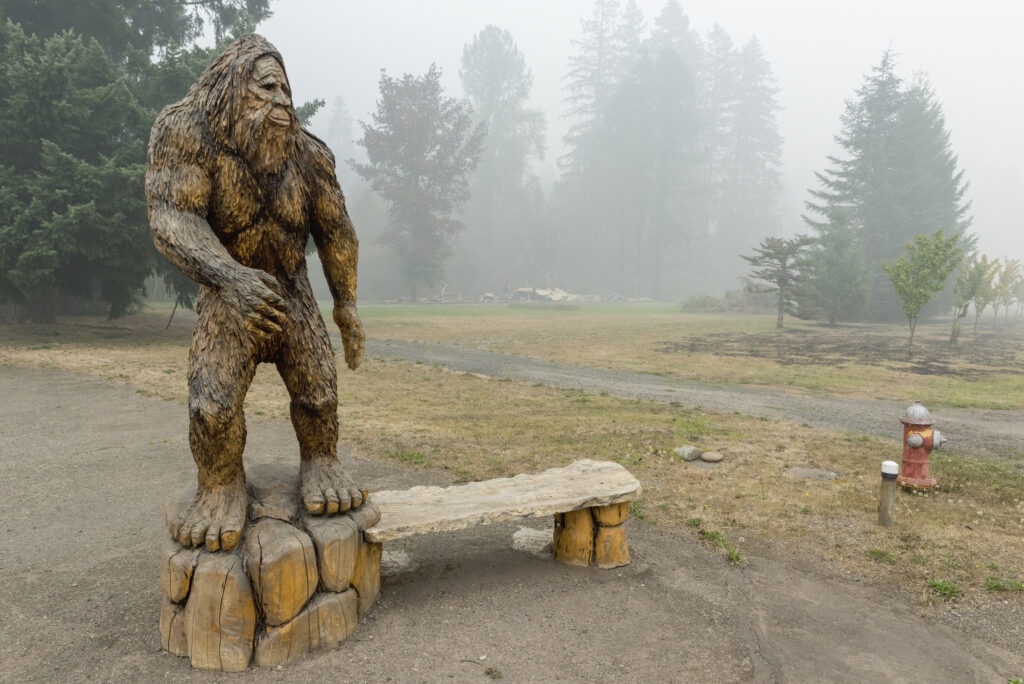 A statue of a large, hairy bipedal figure on rocks stands next to a wooden bench on a clearing in front of a misty forest.
