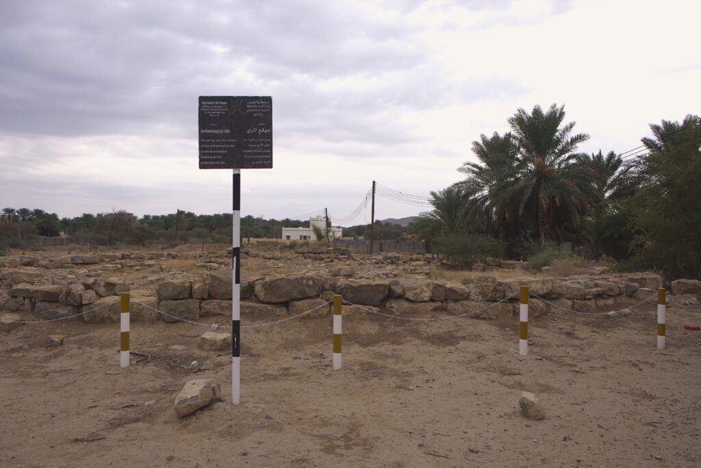 A sign in front of a plot covered in stacked stones reads in English, “Sultanate of Oman—Archaeological site.” The sign also includes other languages.