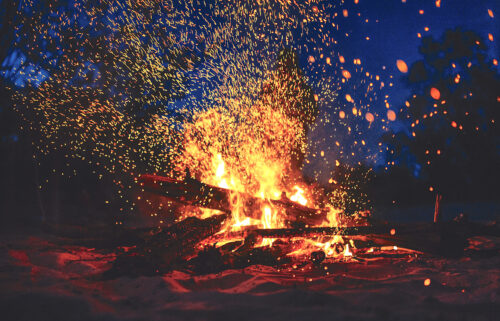 A close-up image features a pile of logs glowing red and surrounded by orange flames. Sparks fly up against a dark blue night sky.