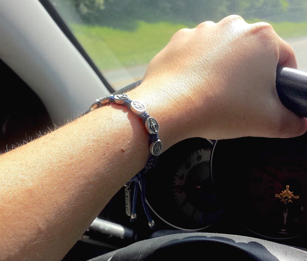 A close-up image features a person’s hand holding a car’s steering wheel. On the driver’s wrist is a silver bracelet of circles with figures etched into each.