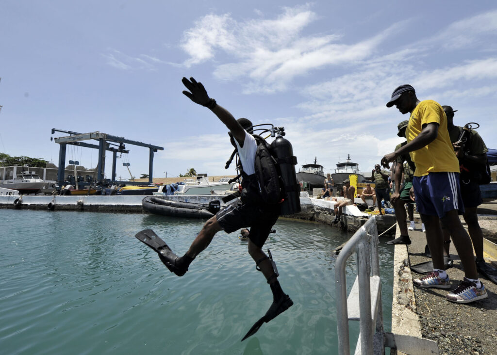 People watch from a stone pier as a person wearing scuba gear, an oxygen tank, and flippers leaps from a metal structure into the adjacent body of water.