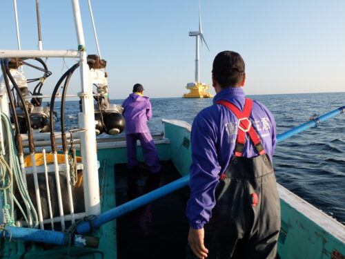 Two people wearing waterproof pants, purple windbreakers, and black caps stand on a teal boat at sea, looking toward a white windmill.