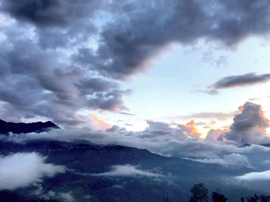 Dark gray clouds hang in a pale blue sky. With orange light shining from behind them, these clouds seem to touch dark rolling hills scattered with trees and buildings.