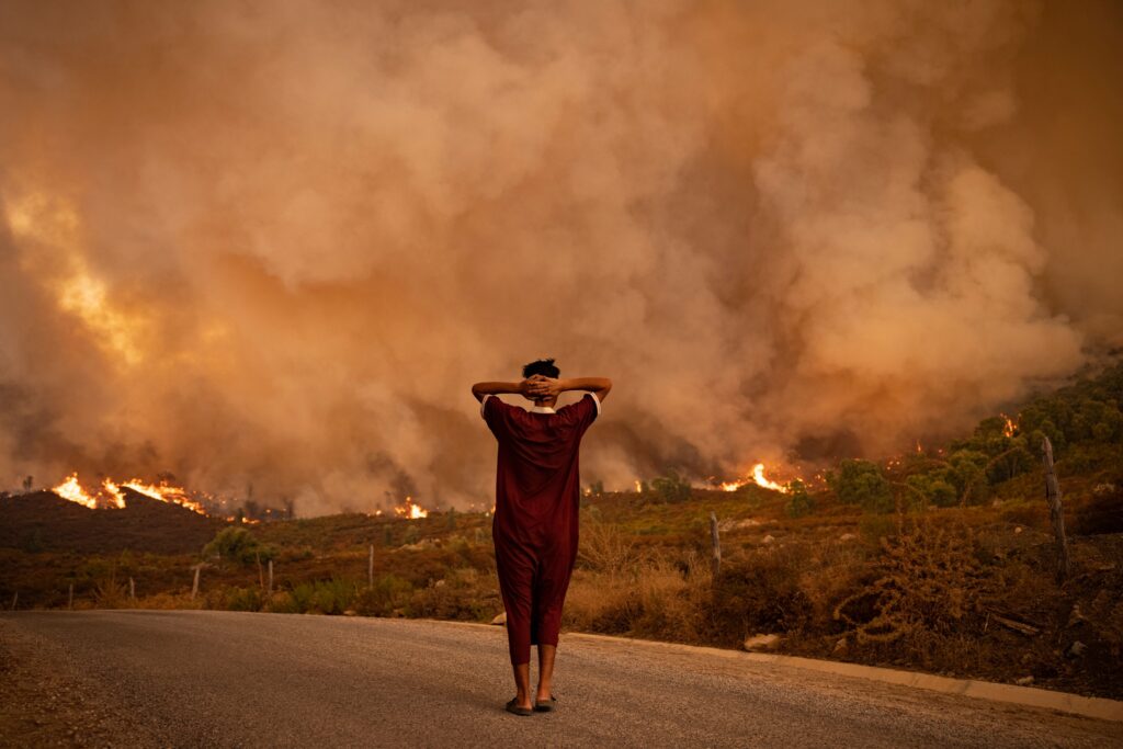 A person in a burgundy jumpsuit stands on a road with their back to the viewer and their hands behind their head. They look into the distance, where yellowed billows of smoke rise from a burning field of grass and trees.