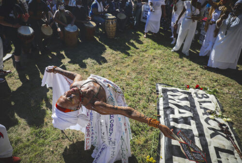 A person wearing a white dress whose skirt is made of ribbons, each with a name written on it, leans back with their eyes closed. They are on grass with a Black Lives Matter banner on the ground beside them and a circle of drummers and bystanders around them.