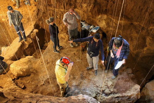 From an aerial view, several people in long-sleeved shirts, khaki pants, and boots are shown standing inside a rocky cave. One person in the center carrying a camera points at two sites, one with each hand.