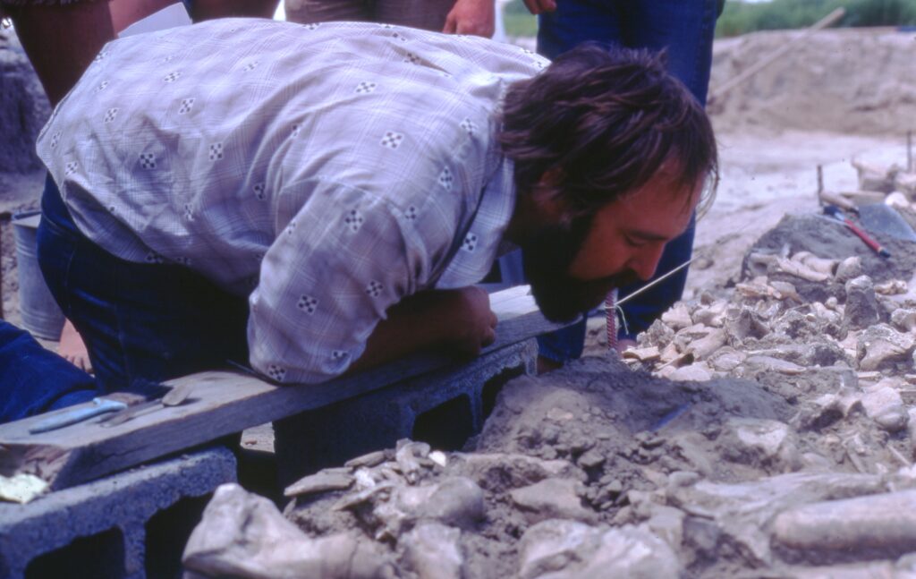 A close-up image features a person with dark hair and a beard in a collared shirt kneeling over an exposed surface in the dirt.