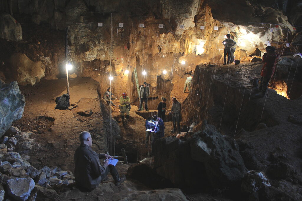 Lit by hanging lights, several people sit or stand inside a cave. Some dig, some write, and some gather in clusters.