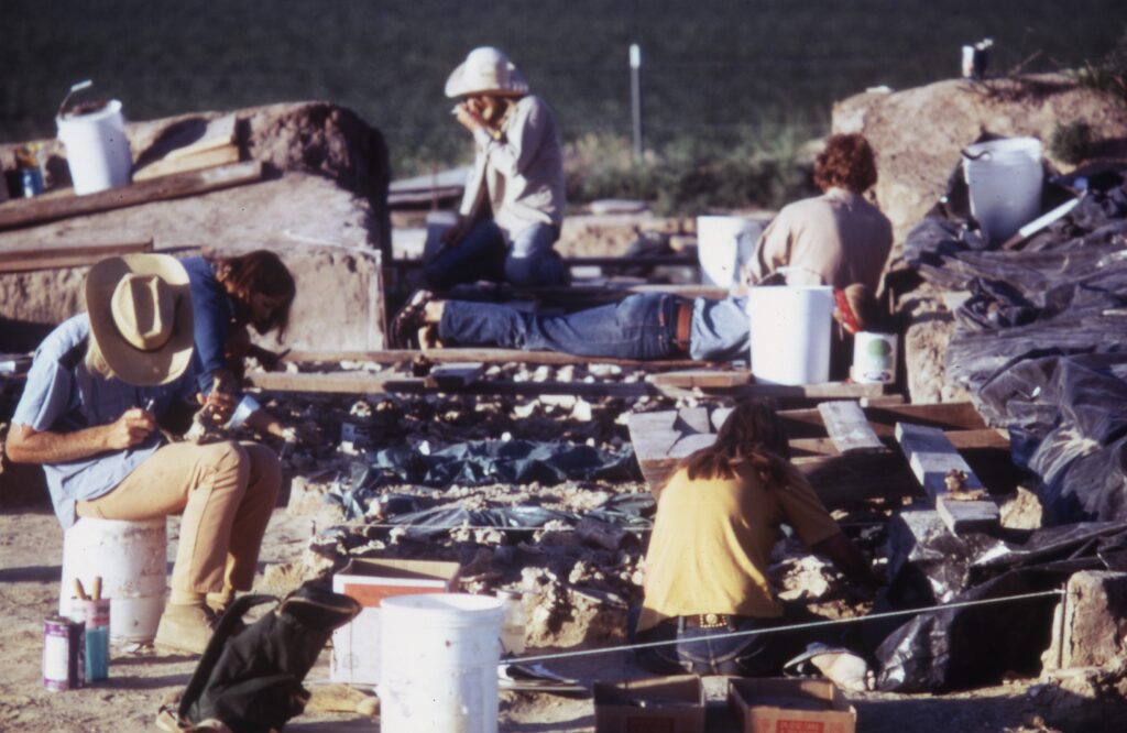 Several people—most wearing brimmed hats, khakis, and work boots—stand or sit near a plot of land covered with buckets, tarps, backpacks, and small objects.