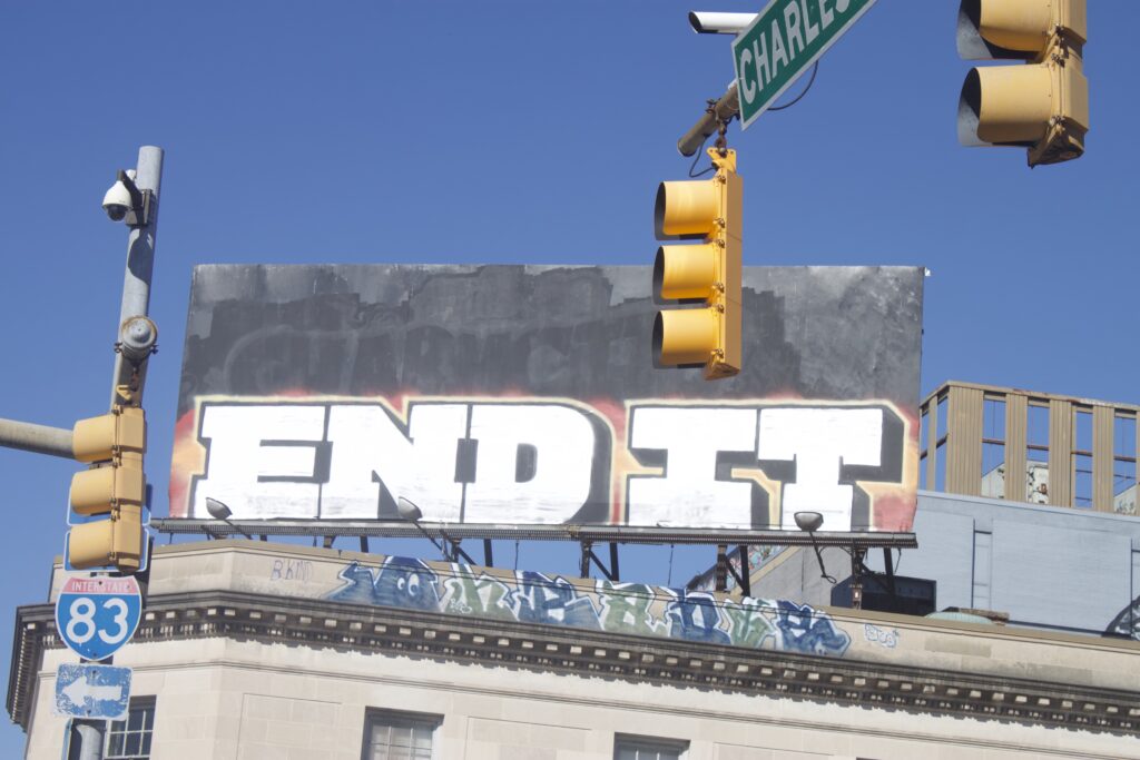 On top of a beige stone building, a giant billboard features bold white text in all capital letters that reads, “End It” against a background that is gray on the top half, and red and yellow on the bottom half.