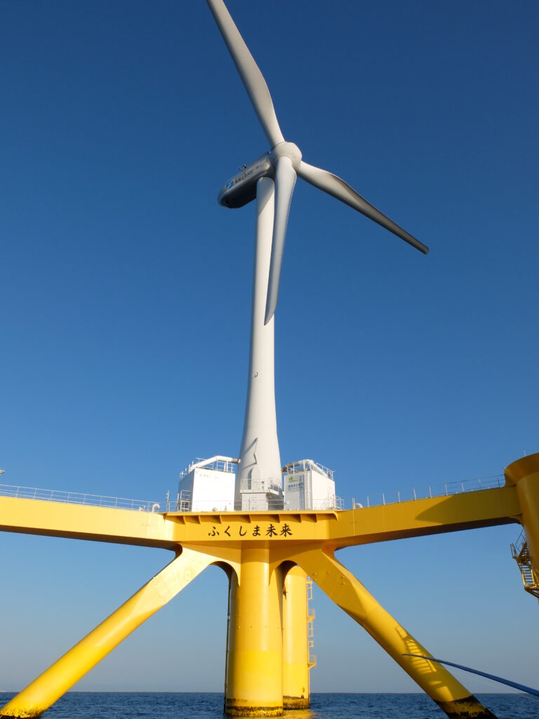 In an open body of water against a blue sky, a large yellow structure holds up an enormous white turbine.