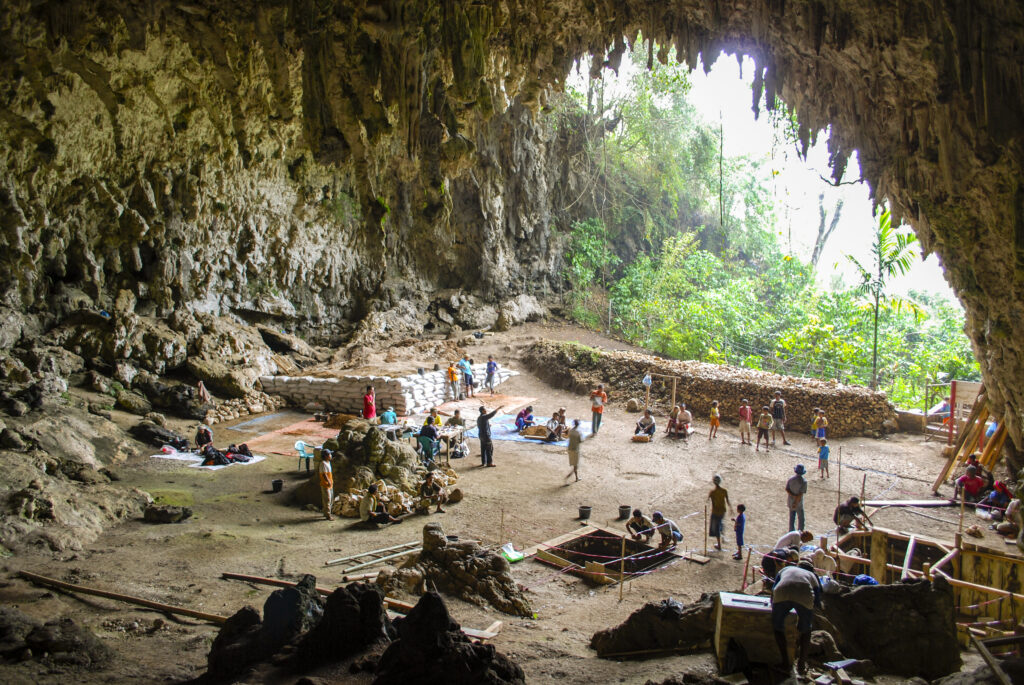 Inside of a large cave, several people stand in clusters around piles of objects and a large square hole in the ground surrounded by red tape. Trees are visible through the cave’s mouth in the background.