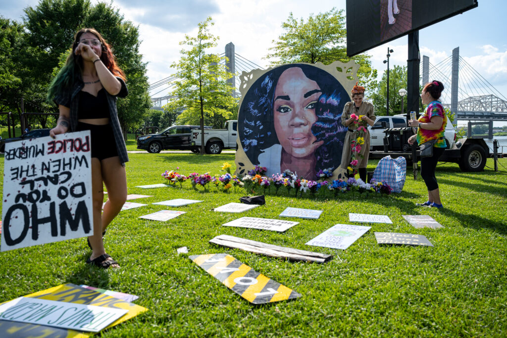 On a green lawn with signs laid across it, two people in the midground hold flowers beside a large circular painting of a woman’s face. In the foreground, a person wipes their eyes and holds a sign that reads, “Who do you call when the police murder?”
