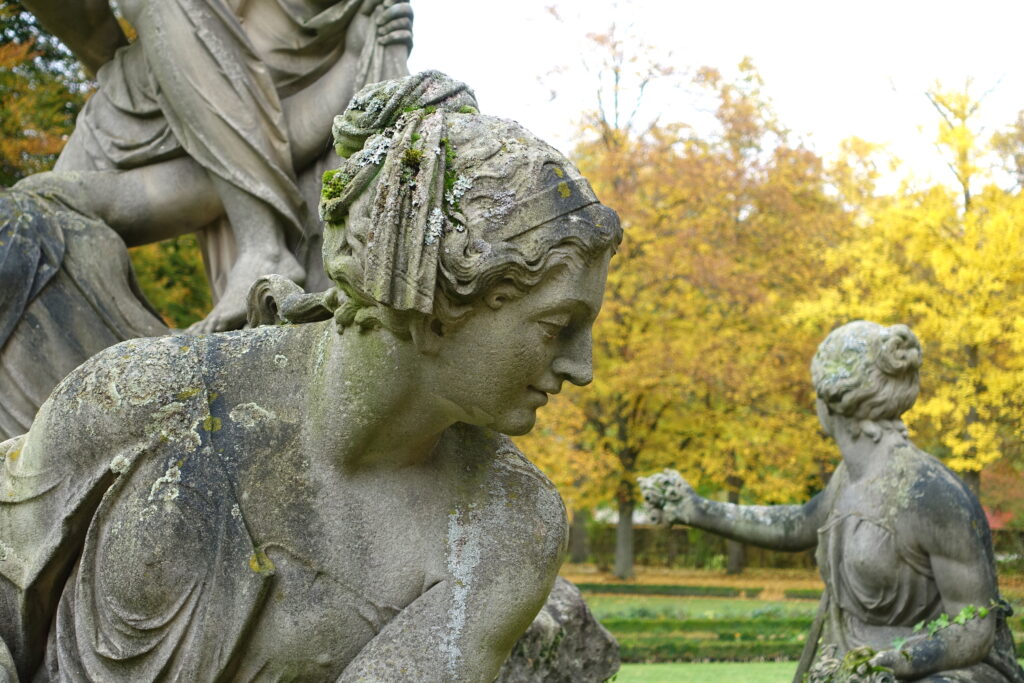In front of green grass and yellow-leafed trees, a close-up image of a statue features a woman’s profile. She wears robes with her hair in a bun and has two other figures, faces unseen, around her.