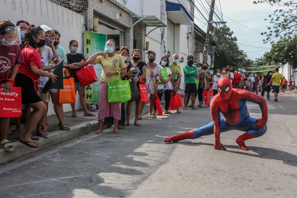 Next to a long line of people wearing clear face shields and blue face masks holding colorful tote bags, a person dressed up in a red and blue outfit with a black spider graphic on its chest area crouches over their left leg with their right fist on the ground.