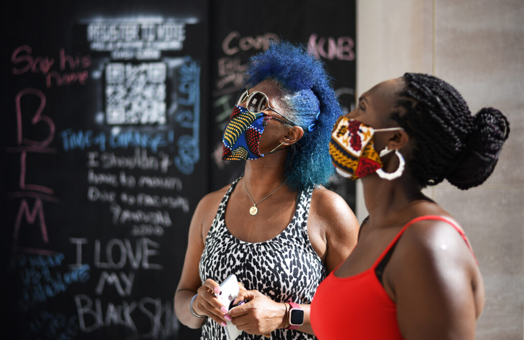 Two people wearing tank tops and colorfully printed face masks look up toward the left corner of the image against a slightly blurred background of a large blackboard featuring various chalk messages. Some read, “BLM,” “I Love My Black Skin,” “ACAB,” and “Register to Vote.”