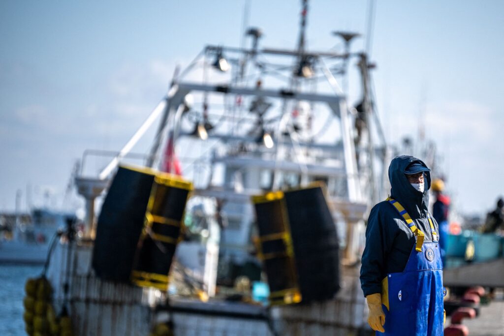 A person wearing blue waterproof overalls, yellow suspenders and gloves, a white mask, and a hoodie and cap stands in front of a slightly blurred background of a docked boat.