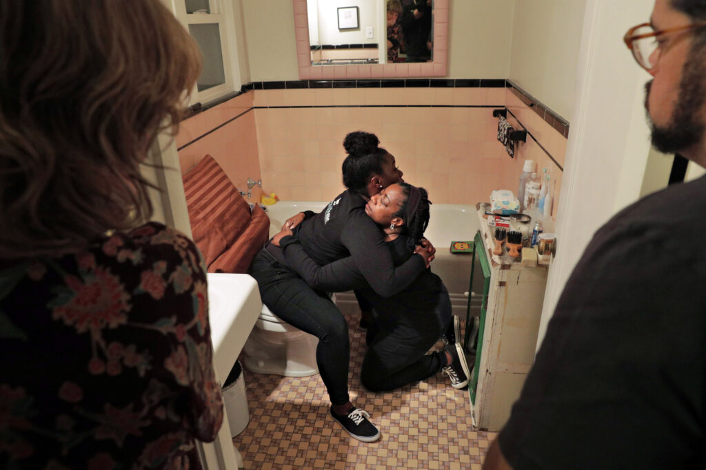 Two people hug in a bathroom, with one seated on a closed toilet and the other kneeling on a tan-and-brown tiled floor. Two standing onlookers flank the image’s foreground.