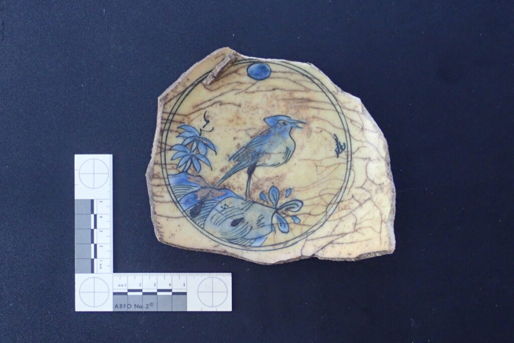 Against a slate blue background, a white, L-shaped ruler sits next to a beige pottery sherd with a blue bird drawn on it.