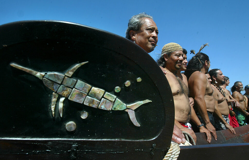 Shirtless people stand in a row with their hands propped on a long wooden object in front of them. In the foreground sits a large black oval-shaped subject with colorful shells arranged on it in the shape of a swordfish.