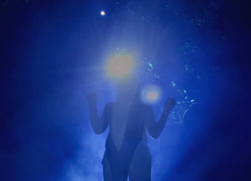 A black silhouette of a person contrasts with an illuminated cobalt-blue background with two bright spotlights shining from behind their head.