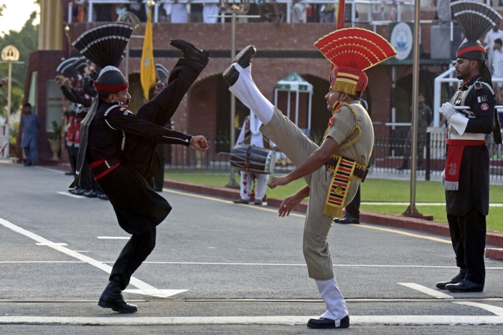 Two people in uniforms—one in black, the other in tan—wearing black boots and hats topped with matching fans face each other and kick one leg up, their feet traveling above their heads. Other people in uniform stand at attention along the roadside behind them.