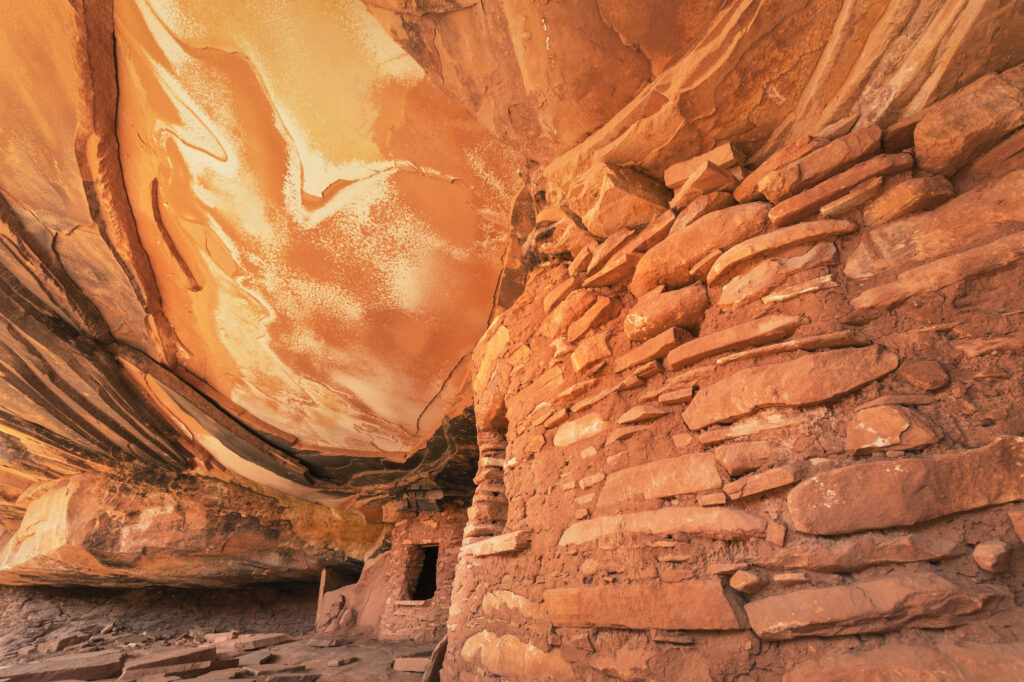 Terracotta-colored structures built of sandstone and mortar have small openings toward the back and middle of the image beneath a sandstone alcove ceiling with a more marbled pattern.